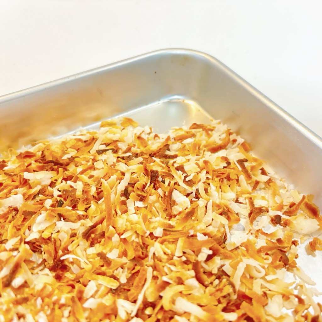The corner of a small, silver pan, spread with a layer of golden, toasted coconut.