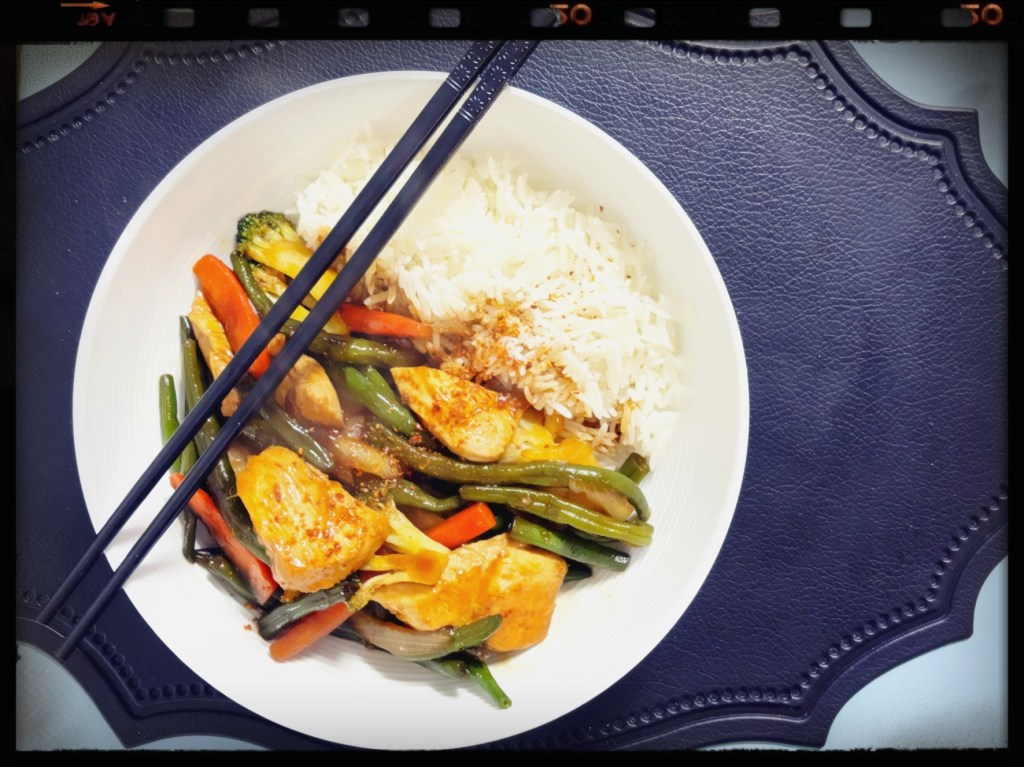A large white bowl, filled half with jasmine rice, half with chicken and vegetable stir fry, and with a pair of chopsticks on the edge of the bowl.