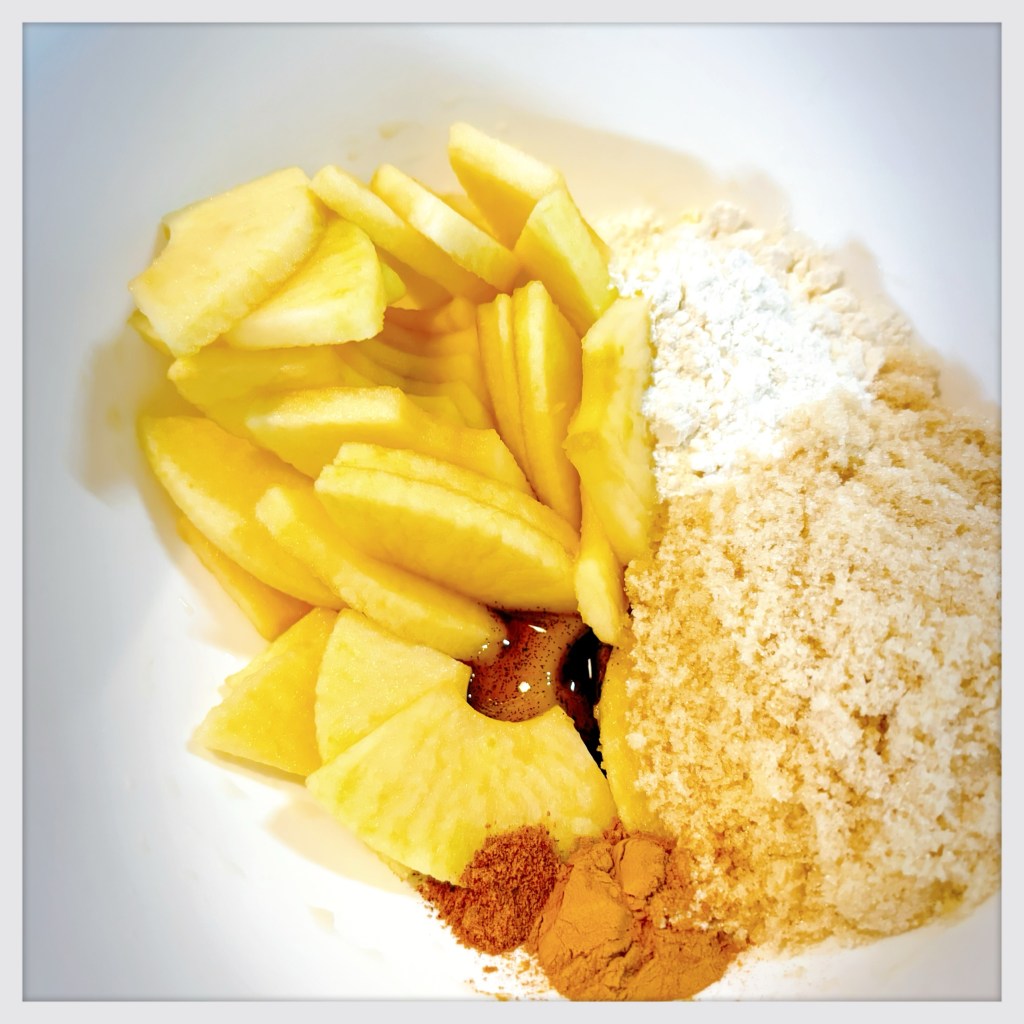A white bowl filled with sliced apples, some flour, some brown sugar, some apple pie spice, and a small blob of vanilla bean paste.