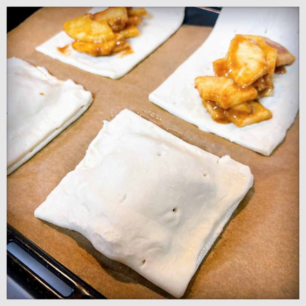 Four sheets of rectangular pastry dough on a baking pan. Two of them are folded simply in half to contain their apple filling, while the other two are still open to illustrate how much can be added to each one, and to still seal it shut without bursting.
