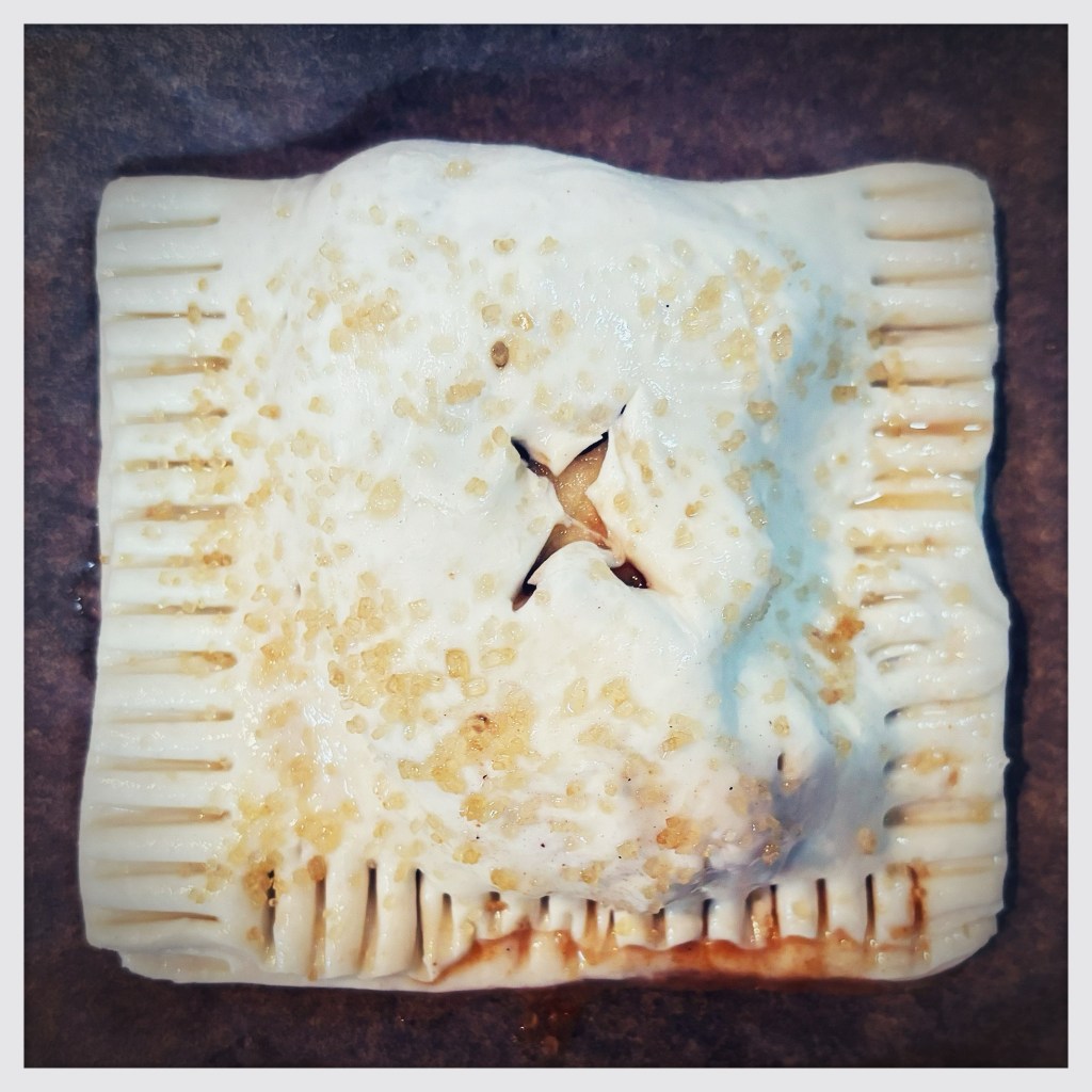 A small, unbaked square of apple pie with fork-crimped edges, a small X carved in the center, and a good amount of crunchy demerara sugar sprinkled on top.
