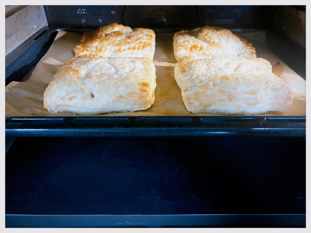 Four small apple pies in the oven. As they are made with a kind of puff pastry, they have poofed up considerably, but they are still too wet and unevenly browned to remove from the oven just yet.