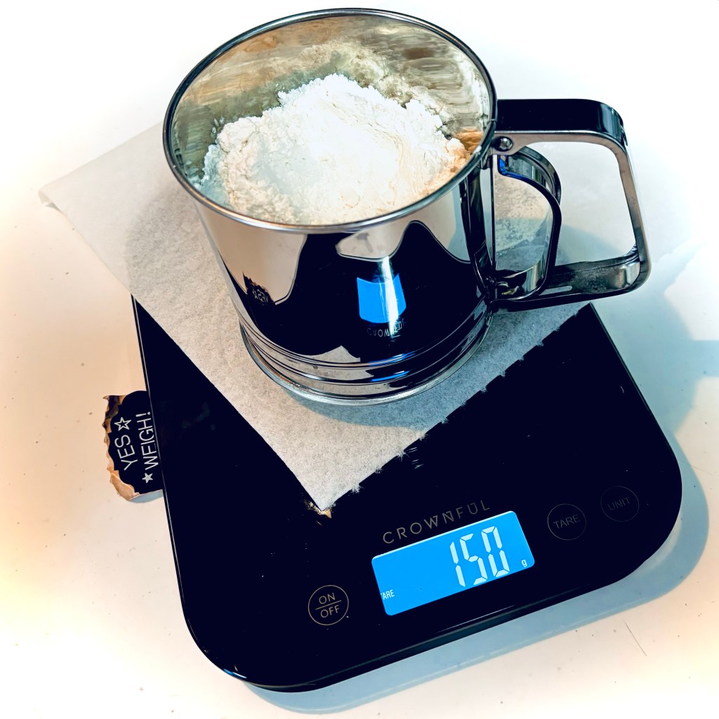 A small metal sifter sits atop a paper towel on an electric scale, which reads 150 grams.