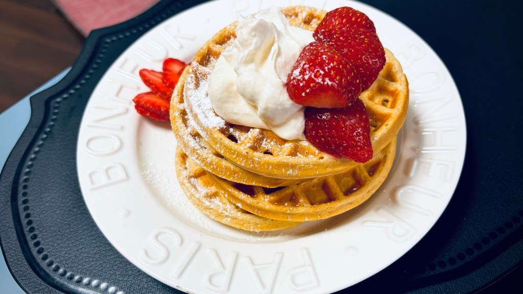 A stack of four small waffles on a white plate, topped with whipped cream, sliced strawberries, and powdered sugar.