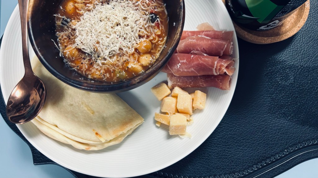 A simple lunch of a bowl of minestrone soup with fresh parmesan grated on top, a simple folded wrap sandwich, some parma ham, and a few small pieces of cheese.