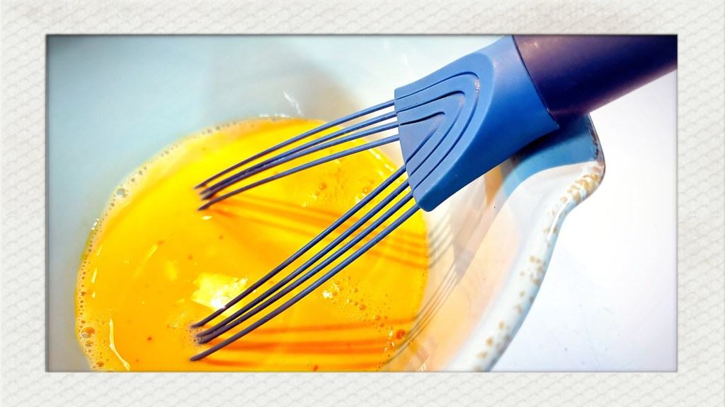 A large, blue ceramic bowl, filled with whipped eggs and a silicone whisk, which is blue too.