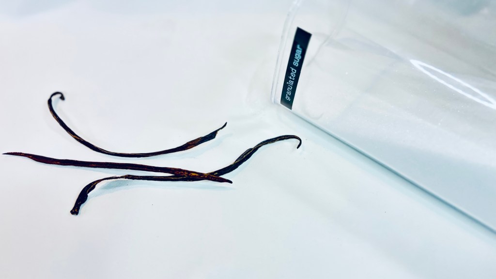 A large, clear container of granulated sugar tipped over in the background, and a few vanilla bean pods in the foreground.