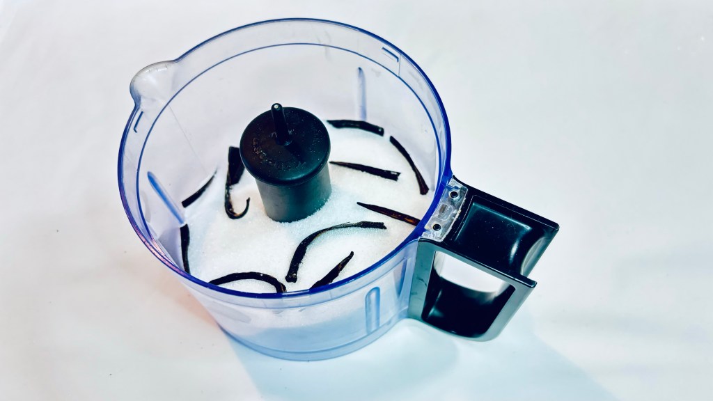 A small food processor bowl filled with some sugar and a number of pieces of broken vanilla beans.