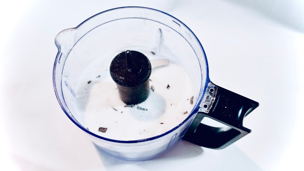 A small food processor bowl filled with some sugar and many itty-bitty pieces of vanilla beans speckled throughout.