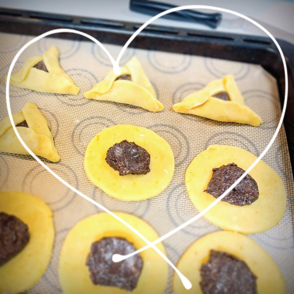 A tray of pastry circles with a mound of dark poppy seed filling in the middle of each, half of them folded into triangular hamantaschen.