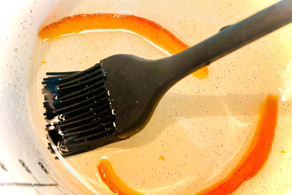 A close up of the inside of a bright white pan, with a small amount of sugar syrup in the bottom of the pot. There are flecks of vanilla beans and pieces of candied orange peel in the syrup, as well as a black pastry brush.