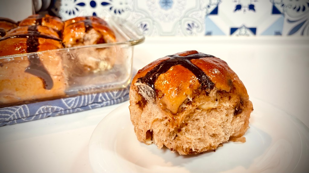A chocolate hot cross bun sits in the foreground, with a panful of more in the background.