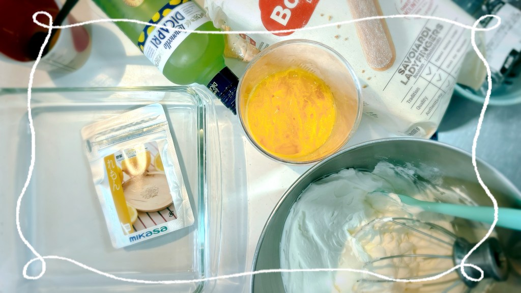 The necessary components of a lemon tiramisu, including a tall glass of lemon curd, a bottle of limoncello, a mixing bowl filled with fluffy mascarpone cream, a bag of store-bought ladyfingers, an empty glass dish, and a small pouch of finely powdered lemon.