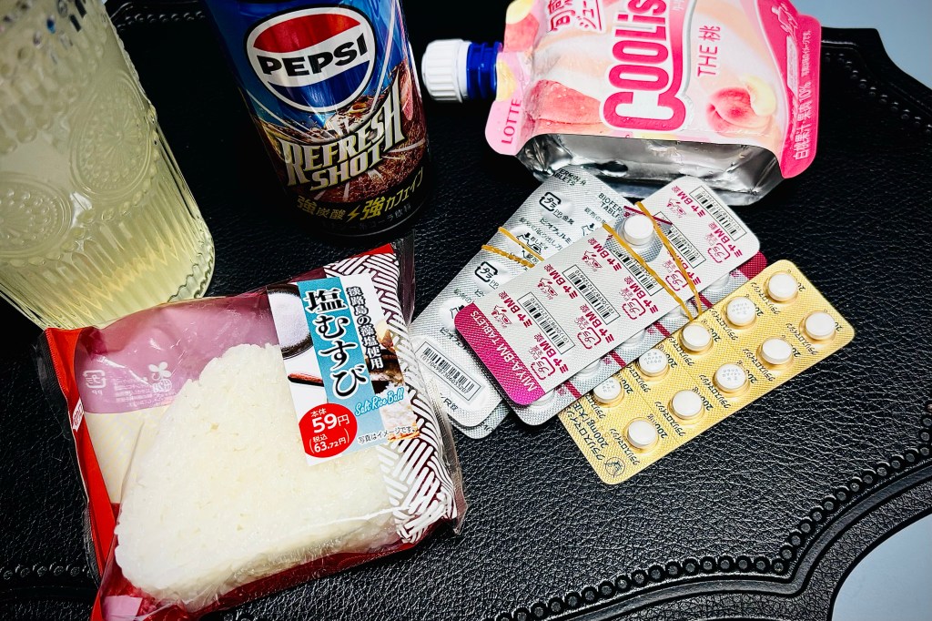 A small table, holding a day's worth of food for someone who is having digestive troubles, including plain rice, a pouch of peach soft serve, a small glass of diluted apple juice, and a wee can of Pepsi. There are also some sheets of antibiotics and probiotics.