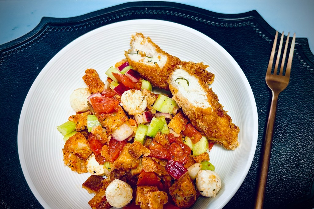 A large white bowl, filled with a meal-size helping of panzanella, with chunks of toasted bread, cut tomatoes and cucumbers and red onions, small balls of fresh mozzarella, and a red wine & herb vinaigrette. There are also two small pieces of fried chicken breast on the side, with a green stripe of shiso leaf in the center of each one.
