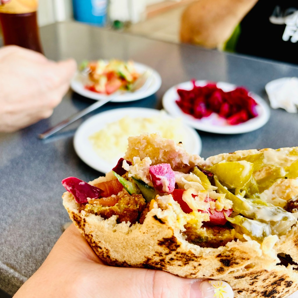 Halfway through a meal of colorful, fresh falafel in a pita, along with hummus, tehina, pickles, tomatoes, and cucumber. In the background are the hands of other people eating, as well as small bowls of salad, pickled cabbage, and beets.